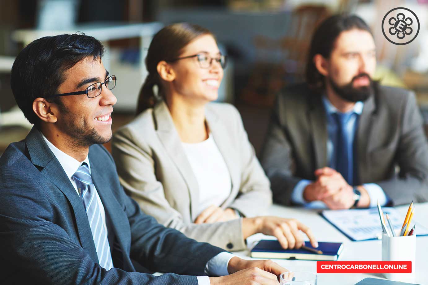Tres profesionales de negocios sonriendo y conversando en una reunión, sentados en una mesa con documentos y una computadora portátil.