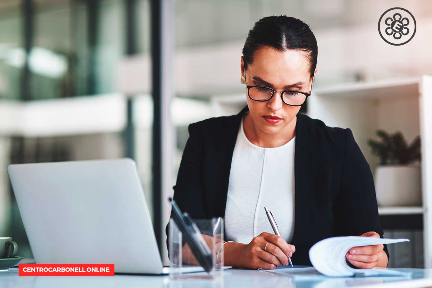 Mujer de negocios con gafas trabajando en un escritorio moderno, escribiendo en un documento y mirando una computadora portátil.