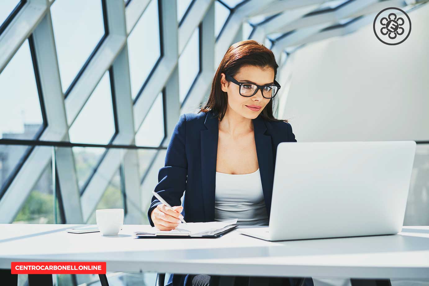 Mujer con gafas de montura negra trabajando en una laptop en un entorno moderno y luminoso.