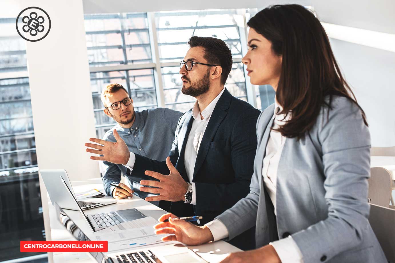 Tres profesionales de la abogacía discutiendo en una reunión, con laptops y documentos sobre la mesa, en un entorno moderno y luminoso.