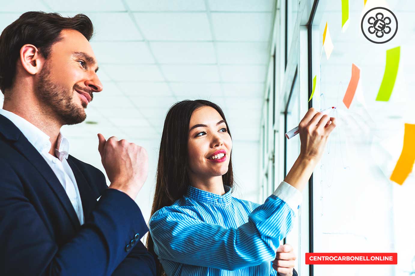 Una mujer sonriente escribe en una pizarra de vidrio mientras un hombre observa, ambos en un entorno de oficina con notas adhesivas de colores.