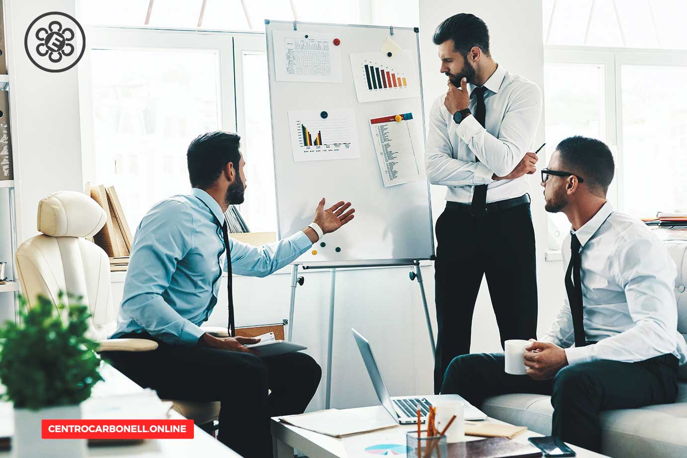 Tres hombres de negocios discutiendo frente a una pizarra en una oficina moderna, con gráficos y datos en presentación.