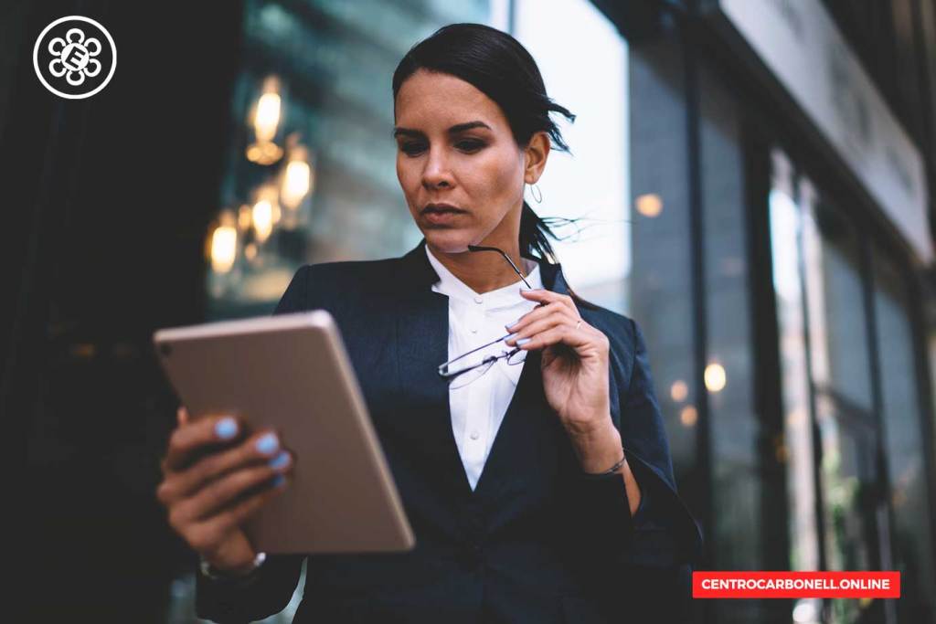 Mujer profesional sosteniendo una tableta y lentes, vestida con un traje oscuro, en un entorno urbano moderno.