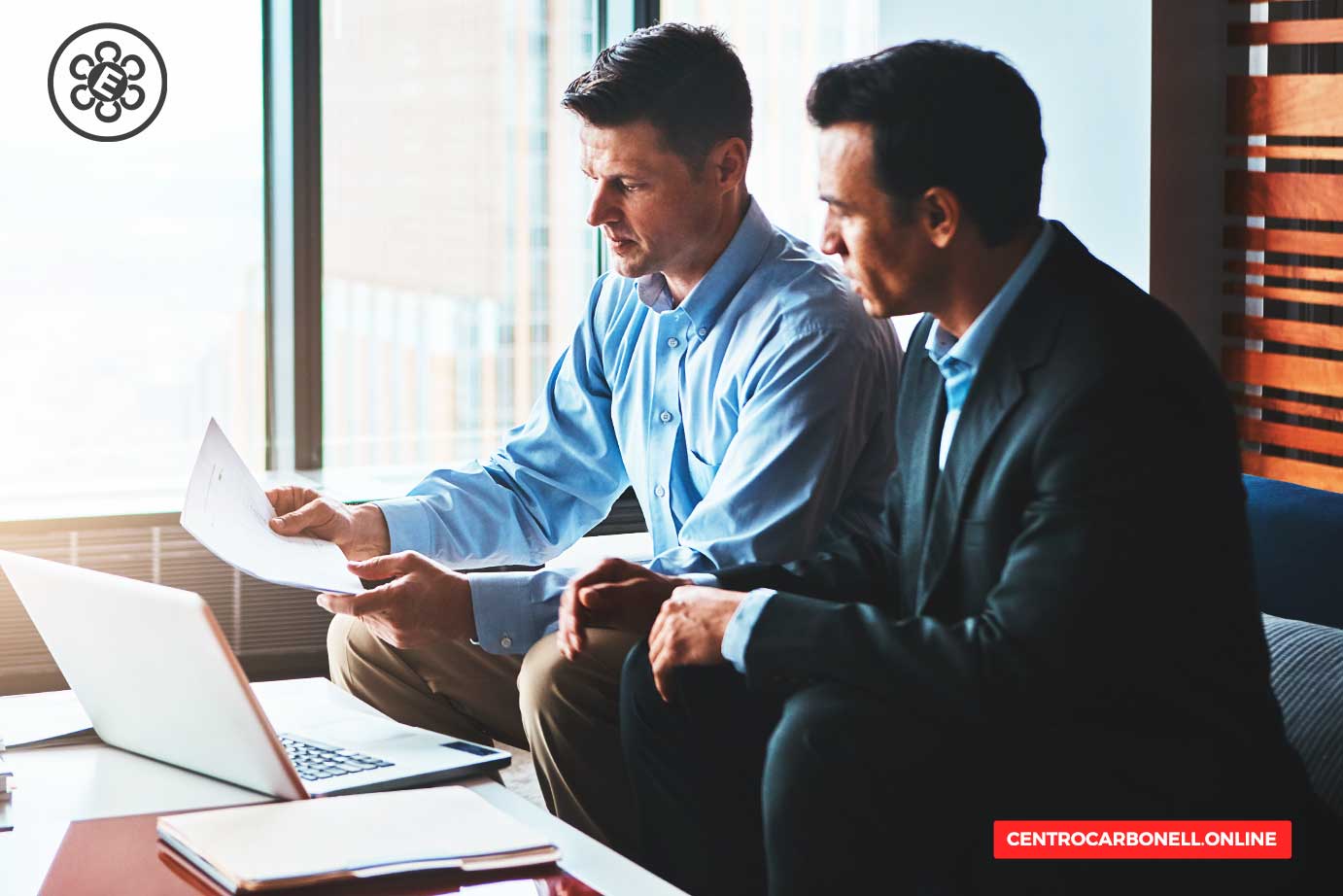 Dos hombres en un entorno de oficina conversando mientras revisan documentos, con una laptop abierta frente a ellos y ventanas al fondo.