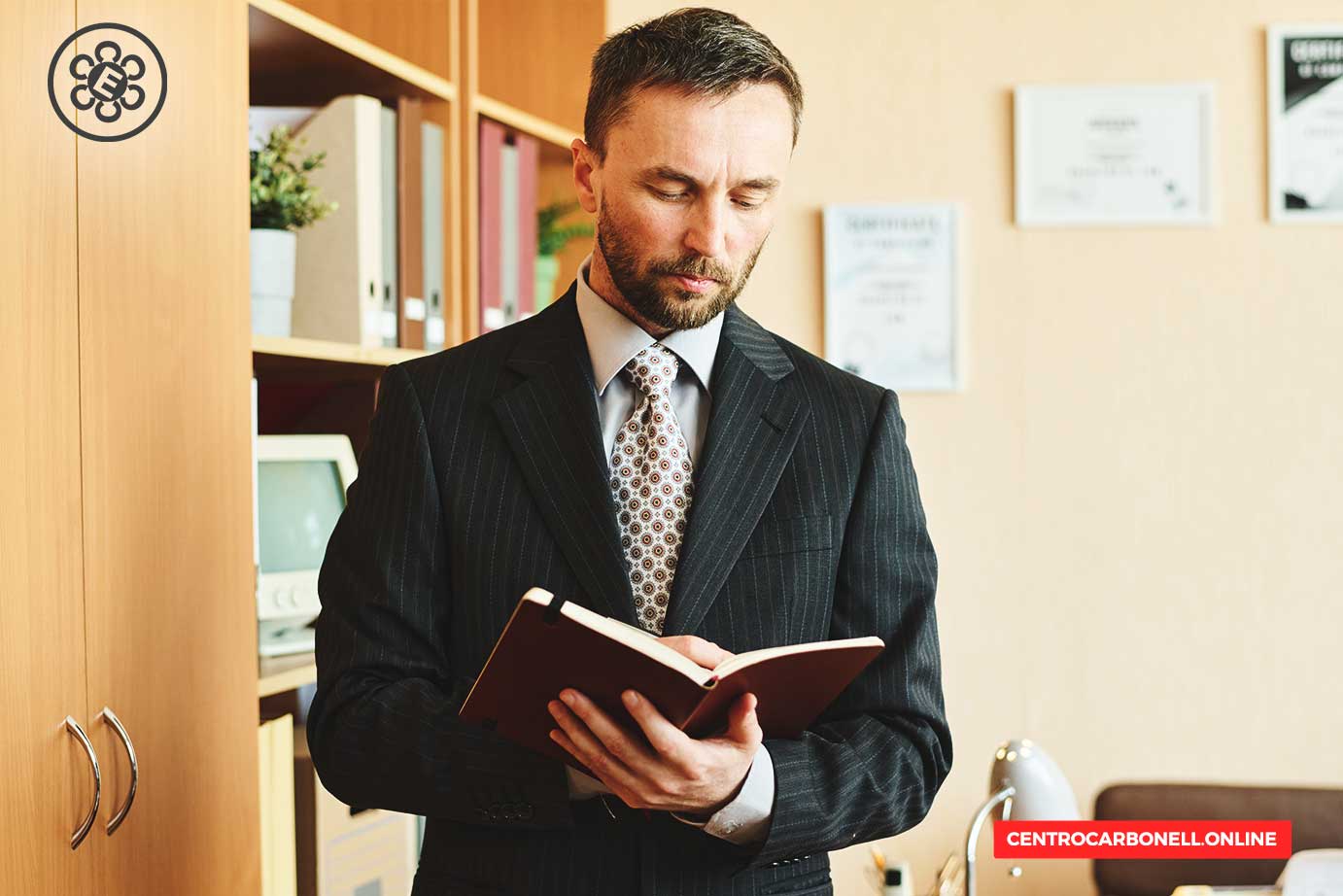 Hombre de negocios con traje, leyendo un libro en una oficina con estantes de archivos y certificados en la pared.