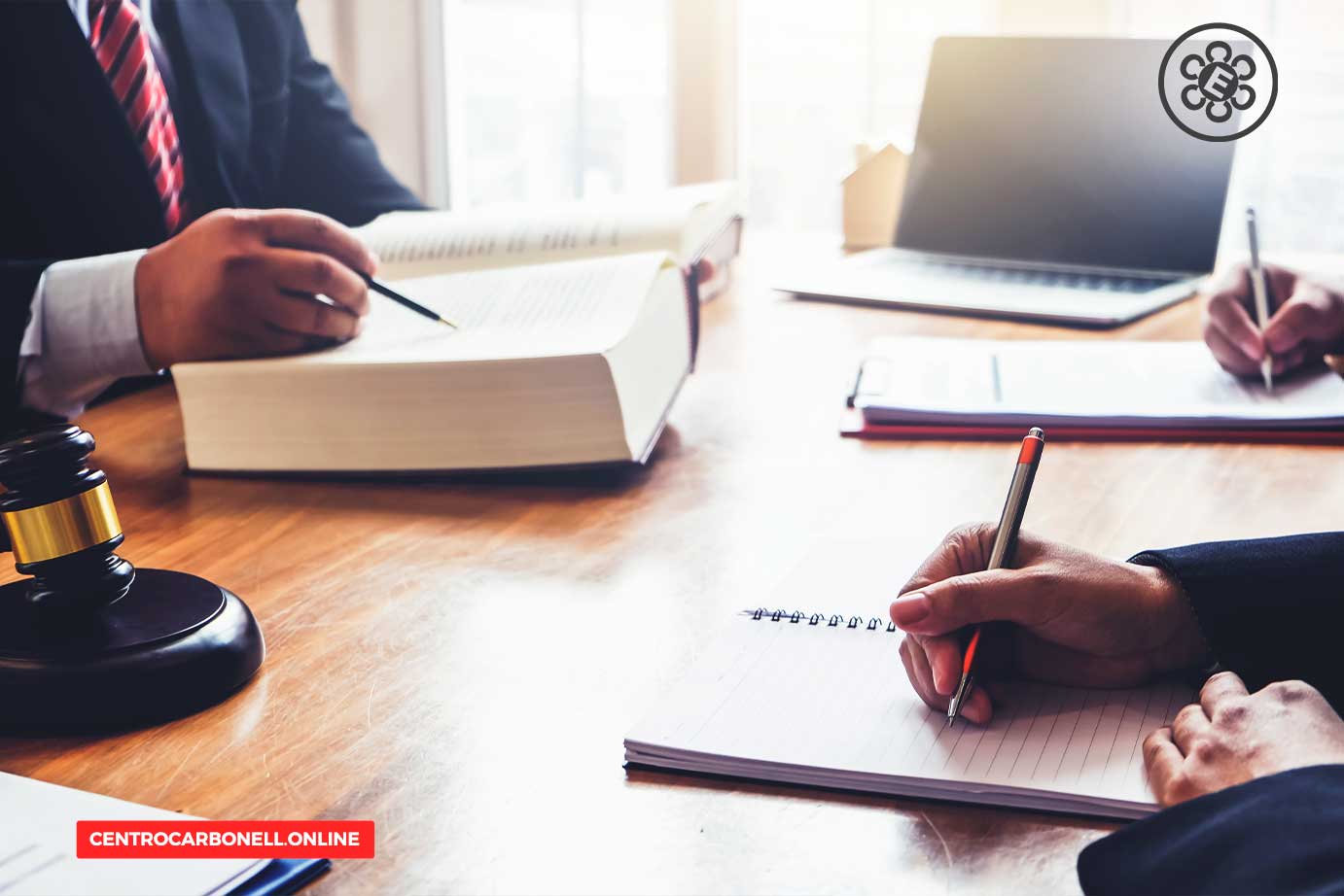 Mesa de trabajo con dos abogados revisando documentos, un libro de leyes abierto y un martillo de juez en el centro.