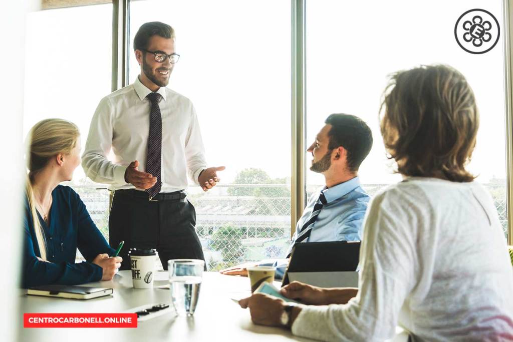 Reunión de negocios en una sala con ventanas, donde un hombre de pie habla mientras cuatro personas lo escuchan atentamente.