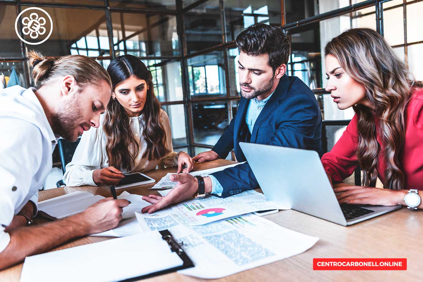 Cuatro profesionales colaborando en una reunión, revisando documentos y gráficos sobre una mesa, con una laptop abierta.