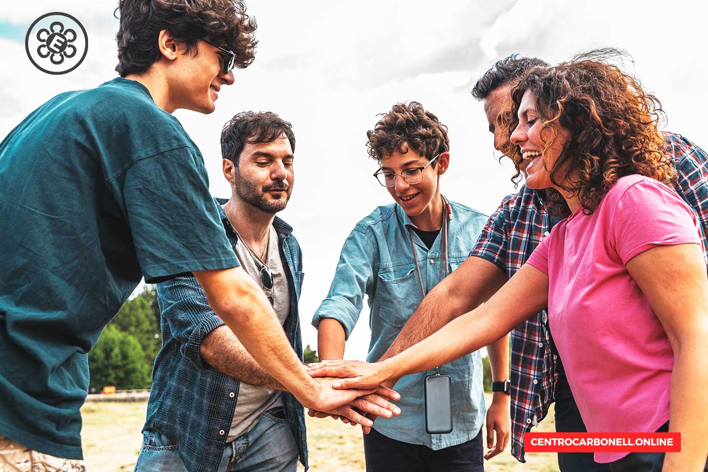 Un grupo de cinco personas sonrientes se unen con las manos en un ambiente al aire libre, en un día parcialmente nublado.