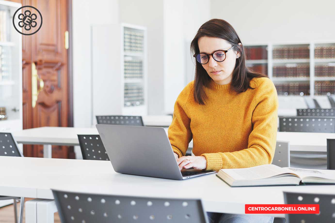 Mujer con suéter amarillo trabajando en una laptop en un aula moderna y luminosa.