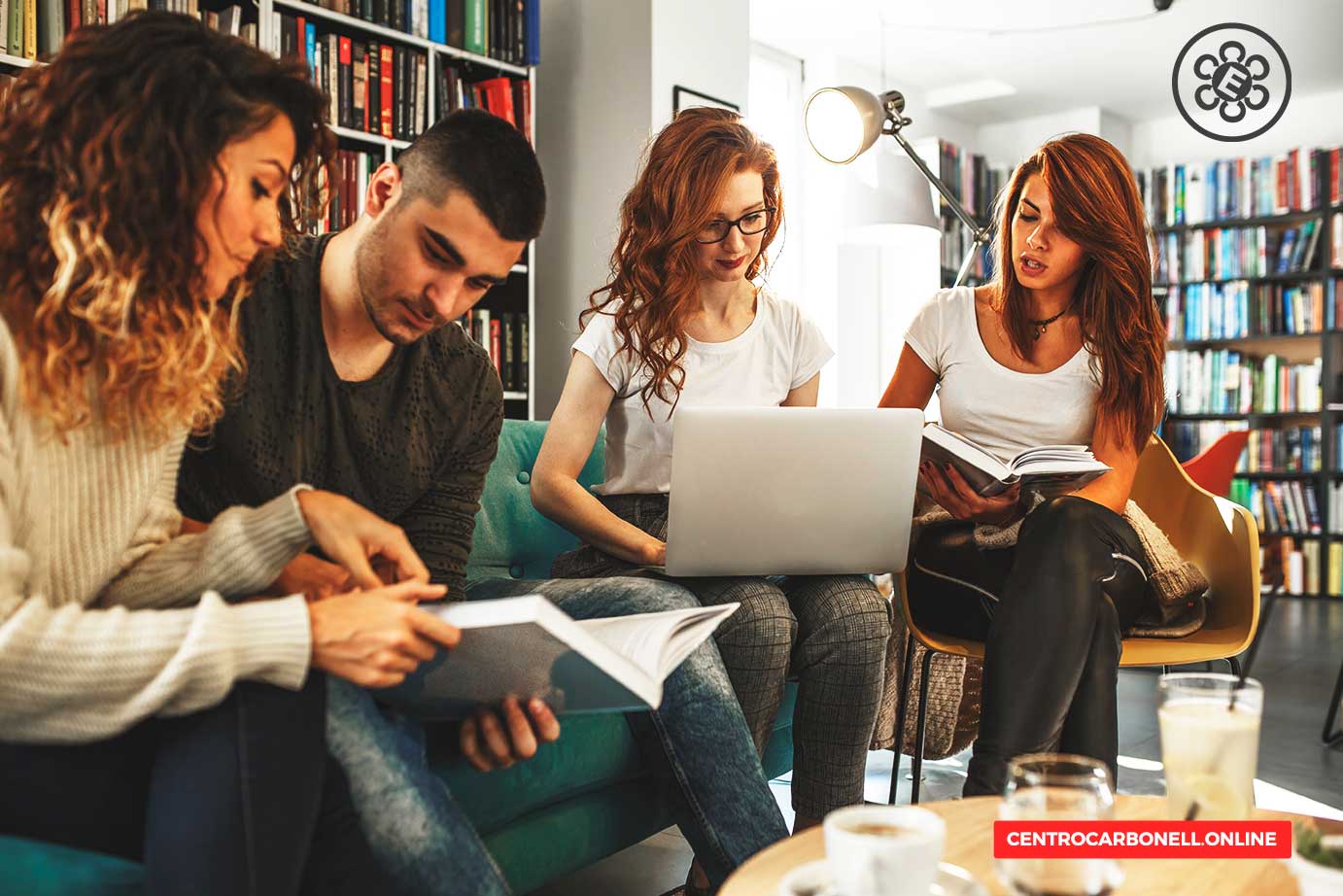 Cuatro jóvenes sentados en un espacio acogedor, revisando libros y utilizando una computadora portátil, con estantes llenos de libros al fondo.
