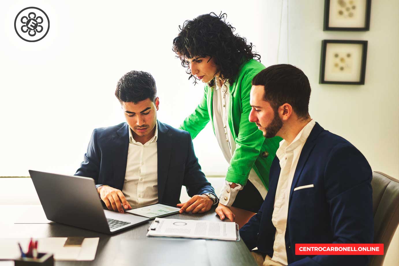 Tres profesionales en una reunión, revisando información en una computadora portátil. Uno de ellos está sentado, mientras una mujer en un blazer verde les da orientación.