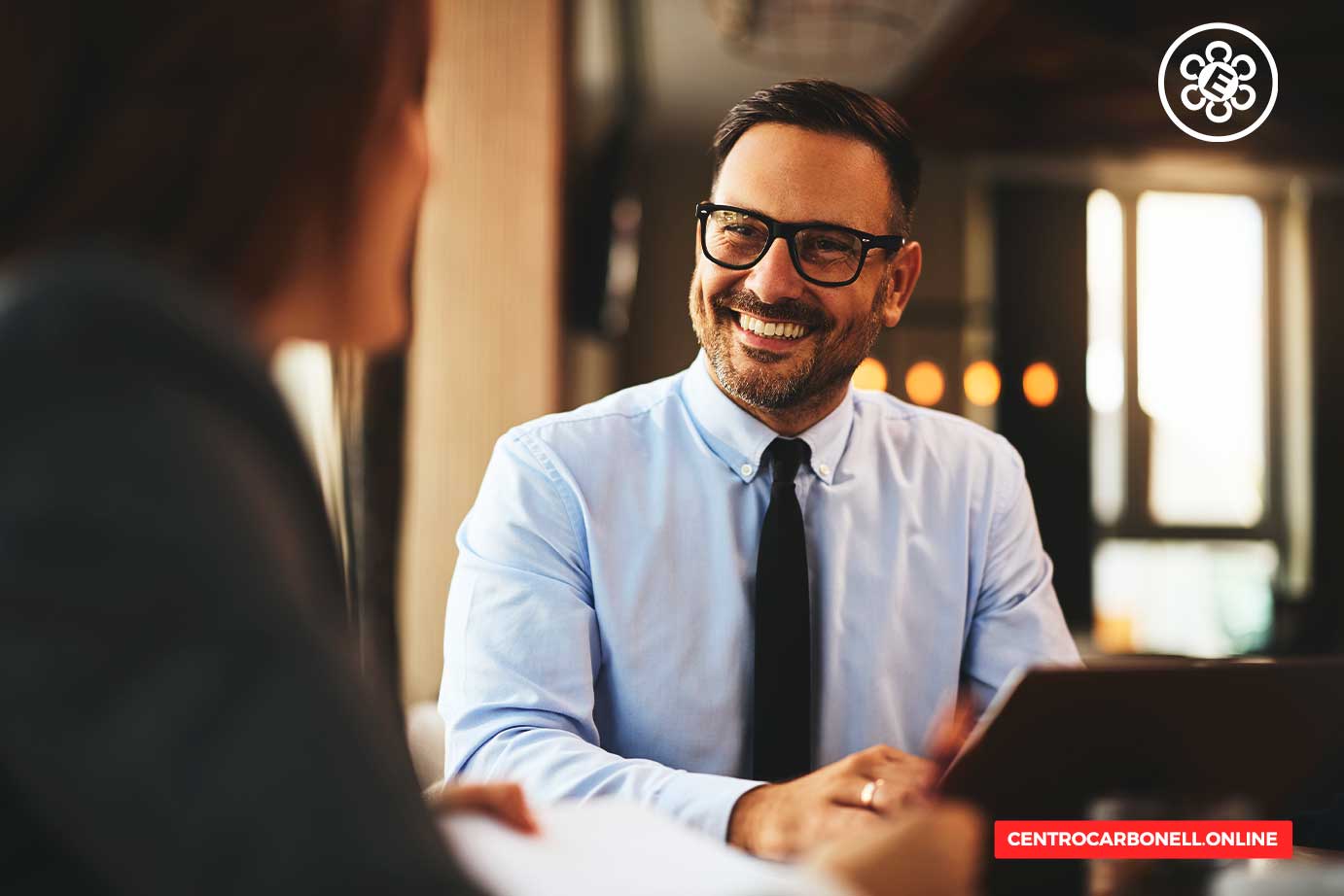 Hombre sonriente con gafas sentado frente a una mujer en un ambiente de oficina, realizando una conversación amistosa.