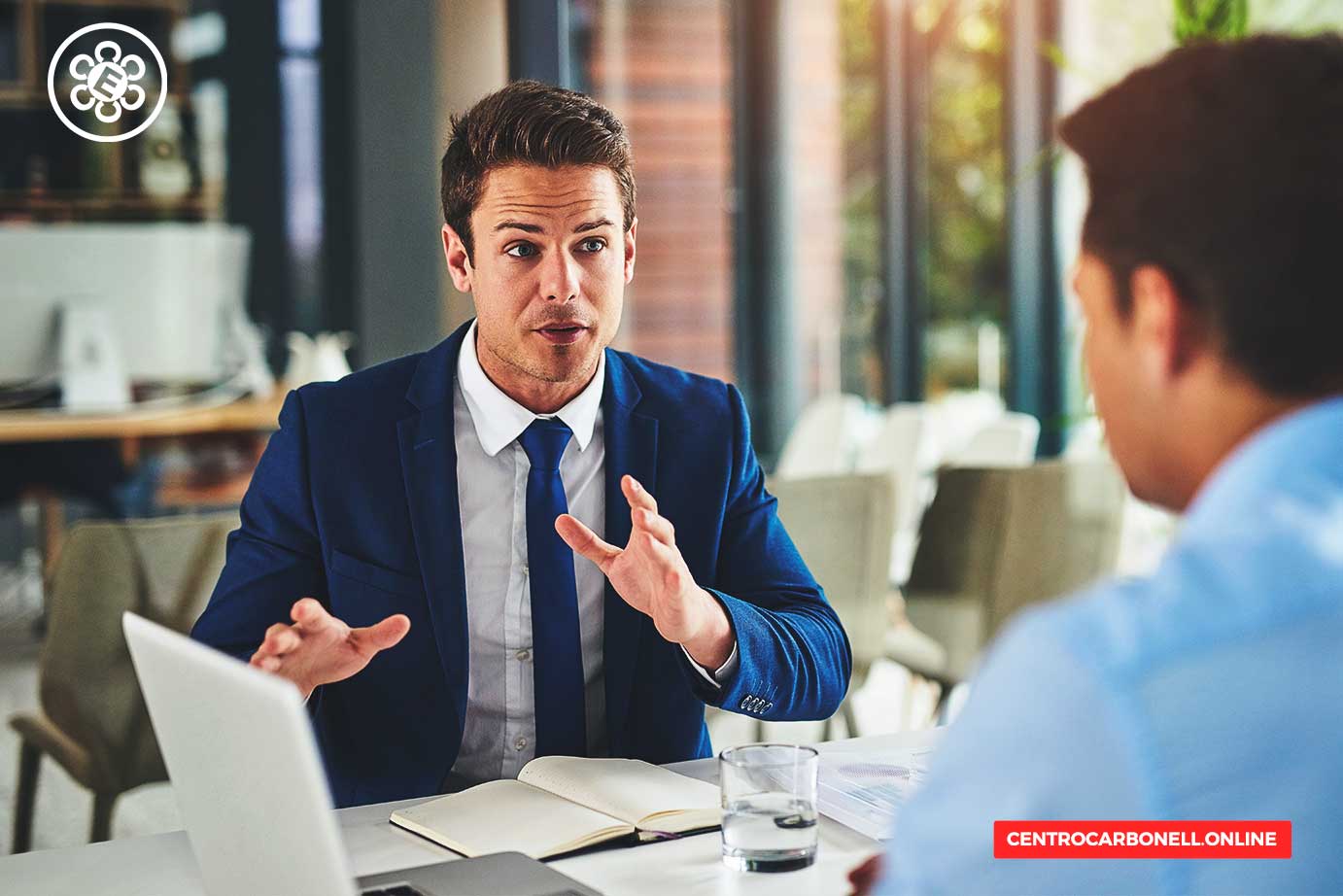 Dos hombres en una reunión de negocios, uno con traje azul hablando con entusiasmo mientras el otro escucha atentamente, frente a una computadora portátil y un cuaderno sobre la mesa.