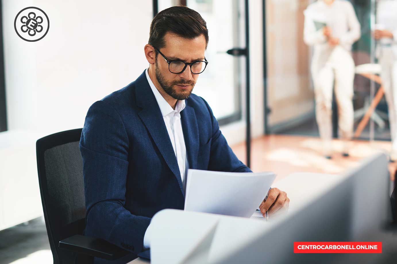 Hombre de negocios con gafas, sentado en un escritorio, revisando documentos en un ambiente de oficina moderna.
