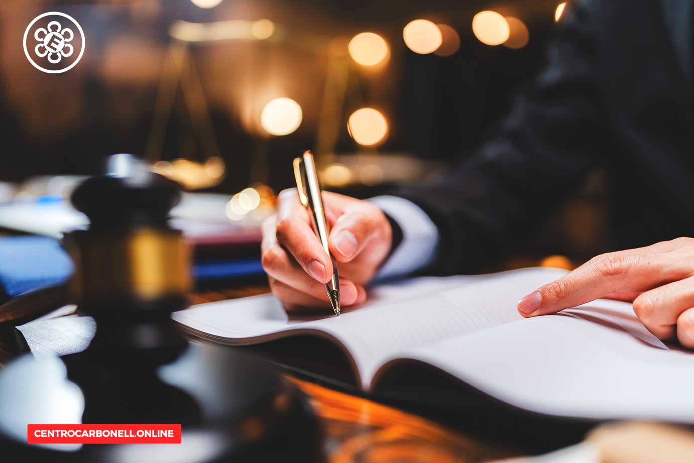 Una mano escribiendo en un cuaderno con una pluma, junto a un mazo de juez en un entorno iluminado con luces suaves.