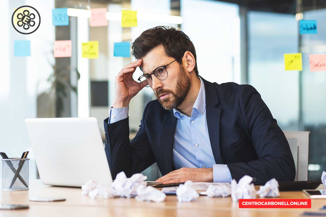 Hombre de negocios con gafas, luciendo pensativo y estresado frente a una computadora portátil, con papeles arrugados en la mesa y notas de colores en el fondo.