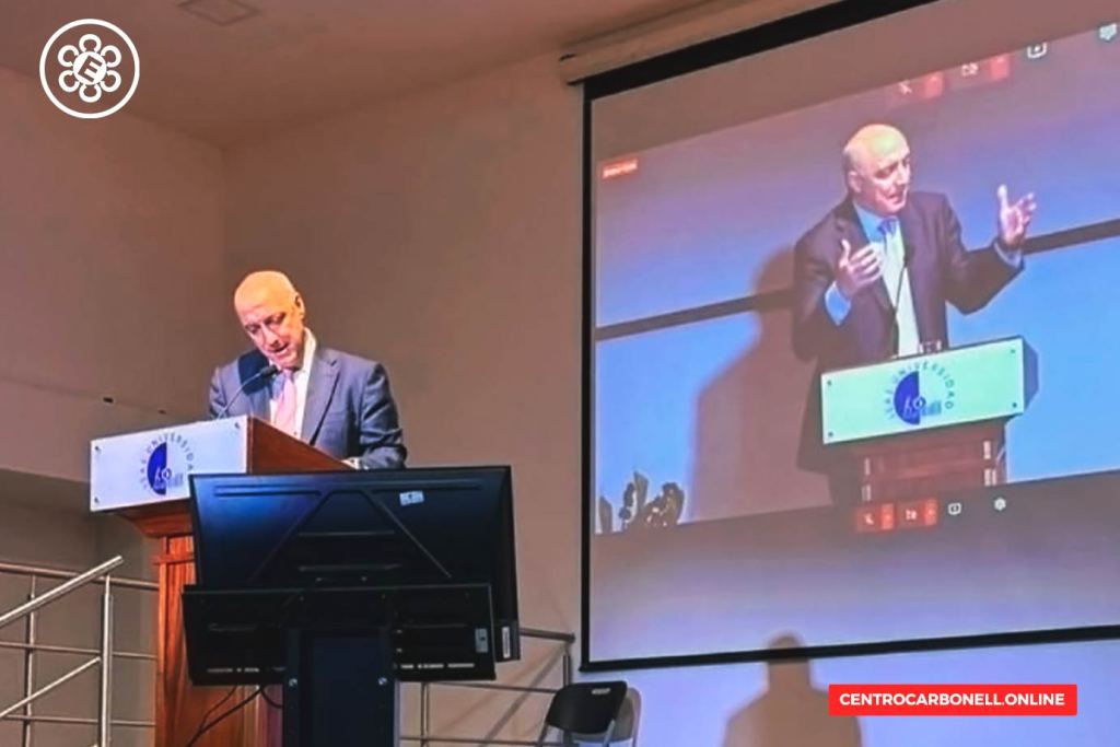 Hombre de pie hablando en un podio durante una conferencia, con una pantalla detrás que muestra su imagen.