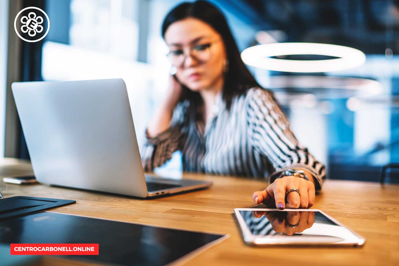 Mujer trabajando en un escritorio con una computadora portátil y una tableta, luciendo pensativa.