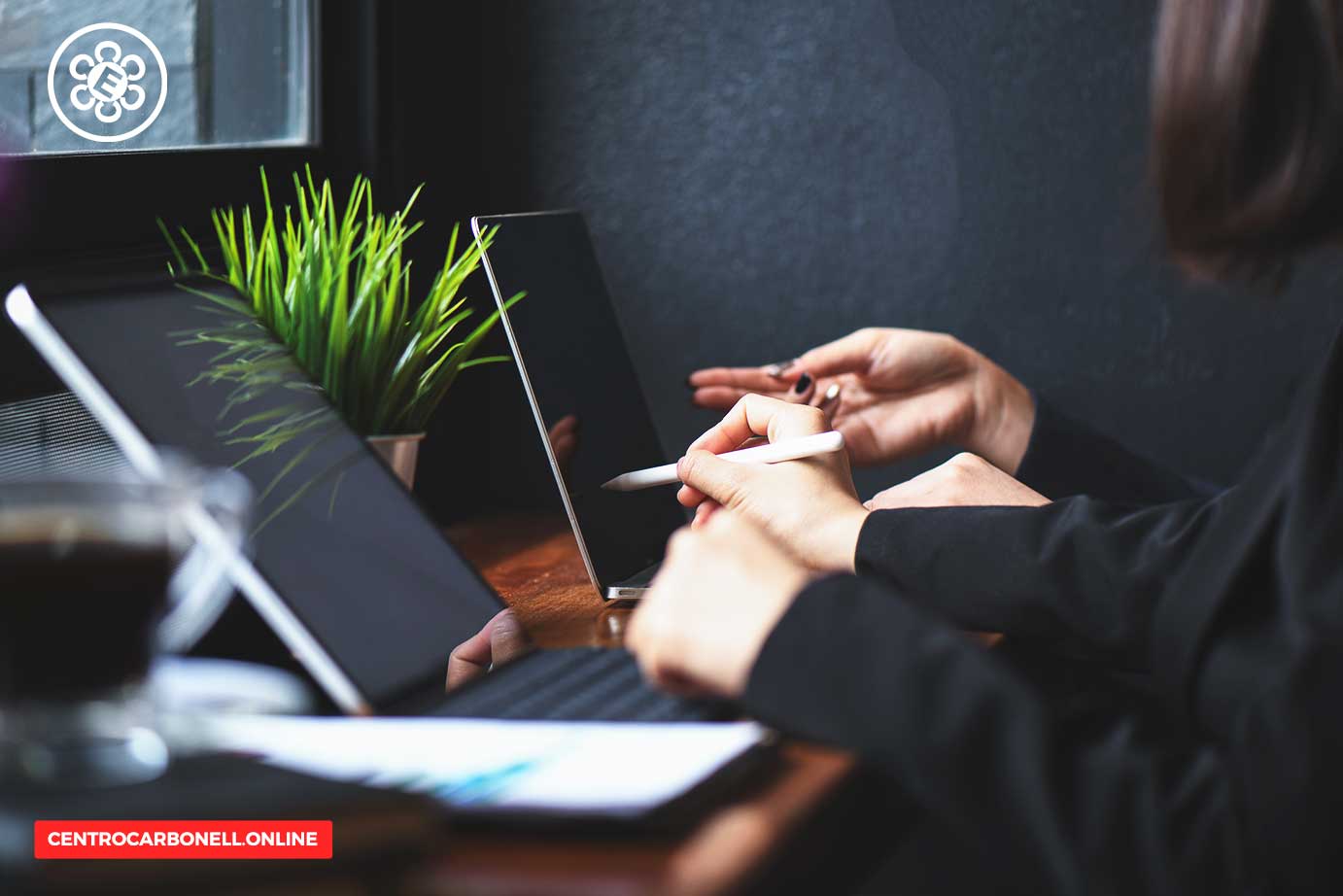 Dos personas trabajando en tablets en una mesa, con una planta decorativa y una taza de café.