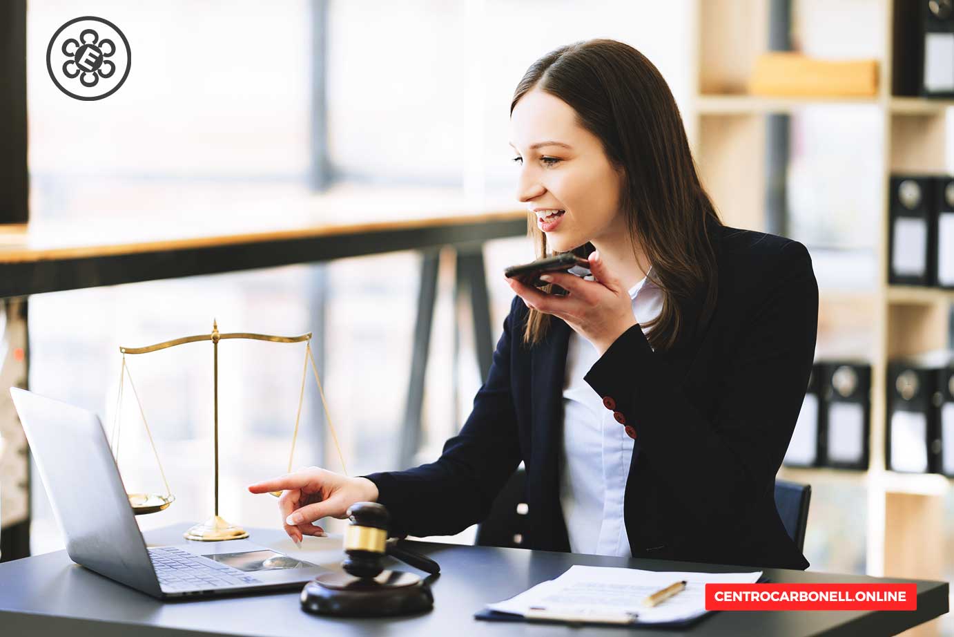 Mujer de negocios hablando por teléfono frente a una computadora portátil en un escritorio, con un libro de leyes y un martillo de juez visibles, y una balanza de la justicia en el fondo.