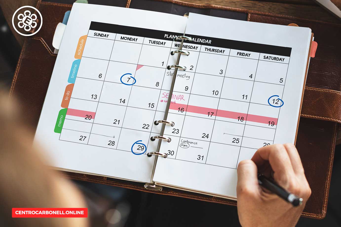 Una persona escribiendo en un calendario planificador abierto con fechas marcadas y eventos destacados, como un seminario y una reunión, sobre una superficie de madera.