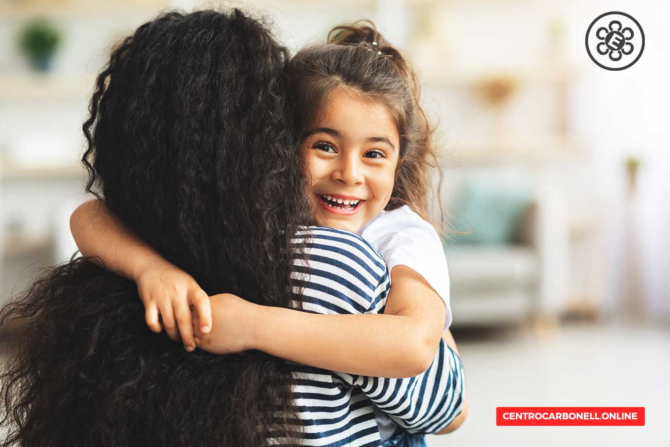 Una niña sonriente abraza a una mujer con cabello rizado detrás de ella, en un ambiente interior acogedor.