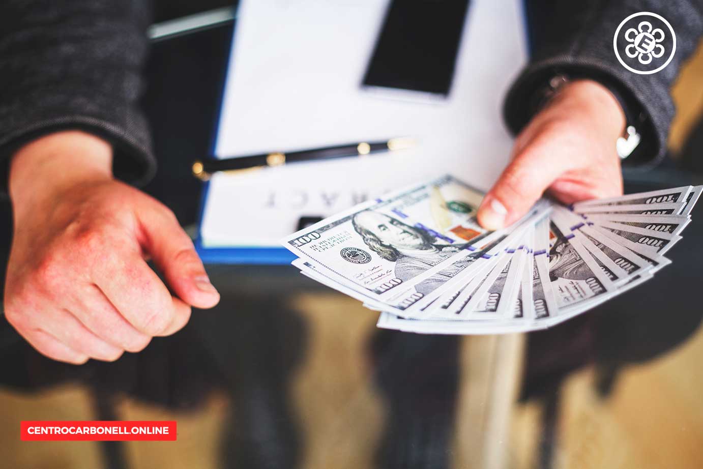 Una mano sostiene varios billetes de 100 dólares, mientras la otra mano descansa sobre una mesa con un bolígrafo y un documento.