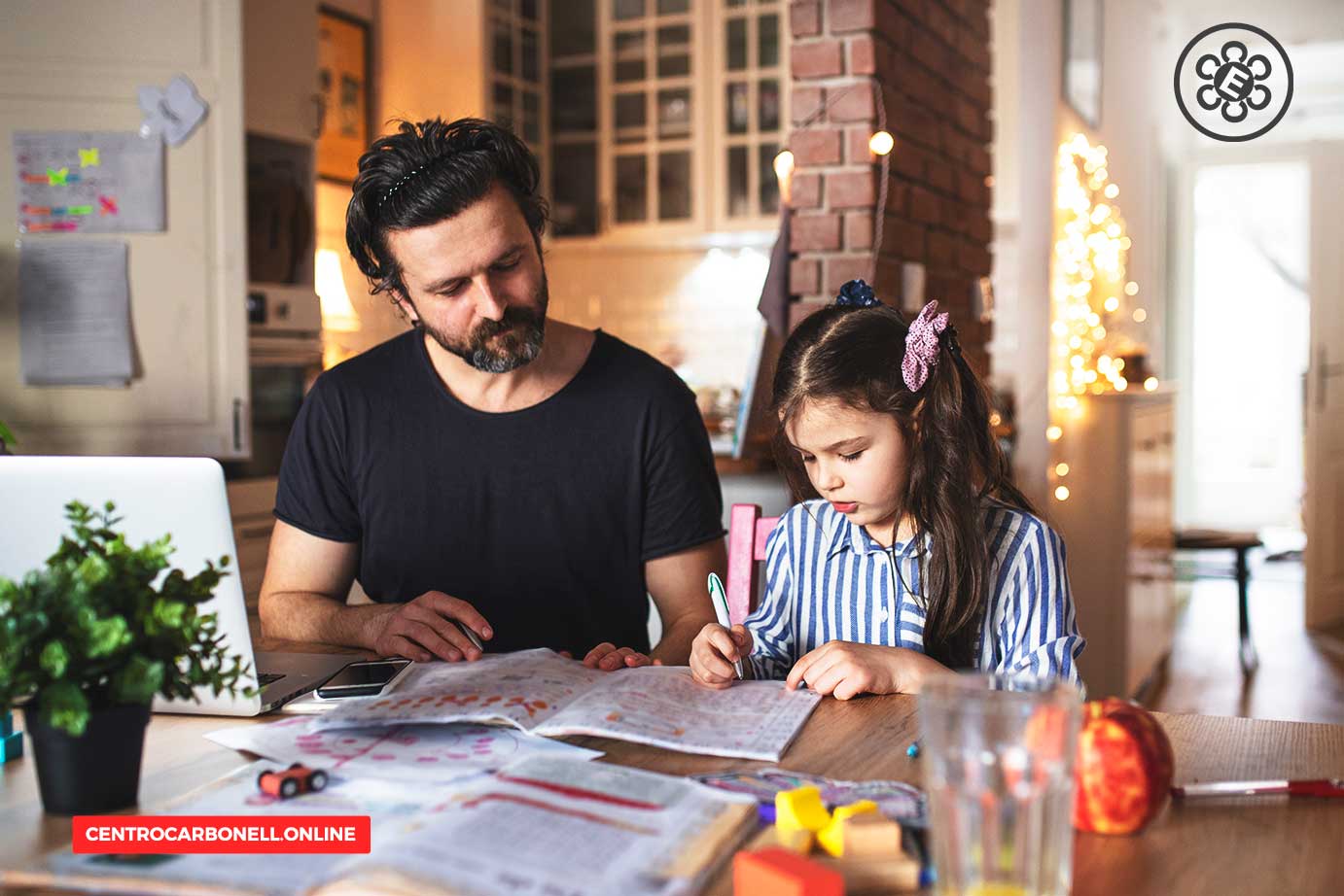 Un padre y su hija pequeña están sentados en una mesa, revisando materiales de estudio. El padre muestra atención mientras la niña escribe. En la mesa hay libros, un vaso, y una planta en una maceta.