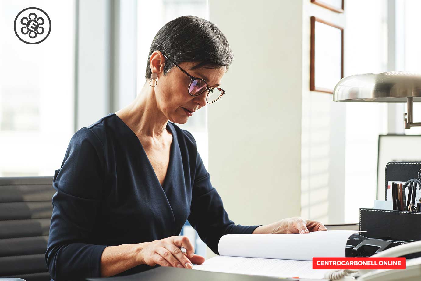Una mujer de mediana edad revisando documentos en su oficina, con una lámpara y material de oficina a su alrededor.