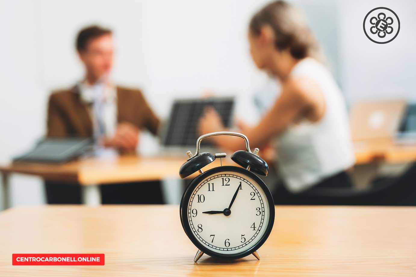 Reloj despertador en primer plano sobre una mesa, con personas trabajando al fondo en una oficina.