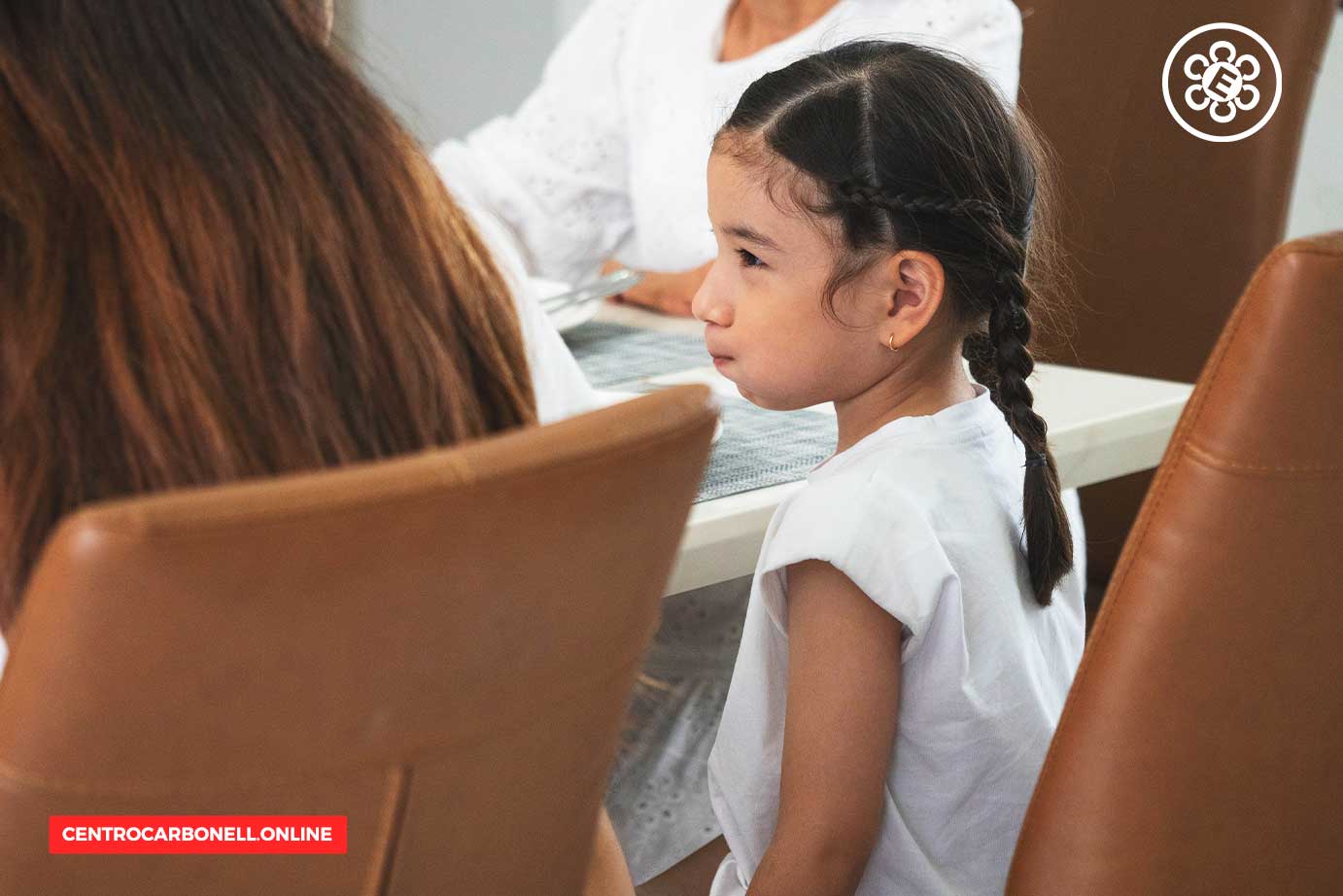 Niña sentada en una mesa, mirando hacia un lado, con cabello trenzado y vestida con una camiseta blanca.