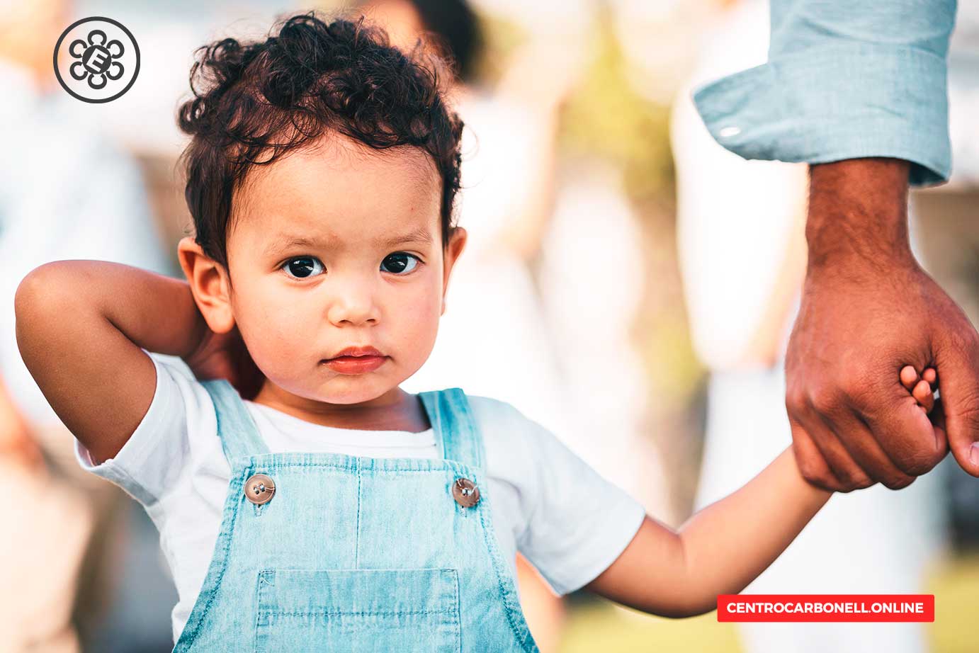 Niño pequeño sosteniendo la mano de un adulto, con una expresión pensativa, vestido con overol de mezclilla y camiseta blanca.