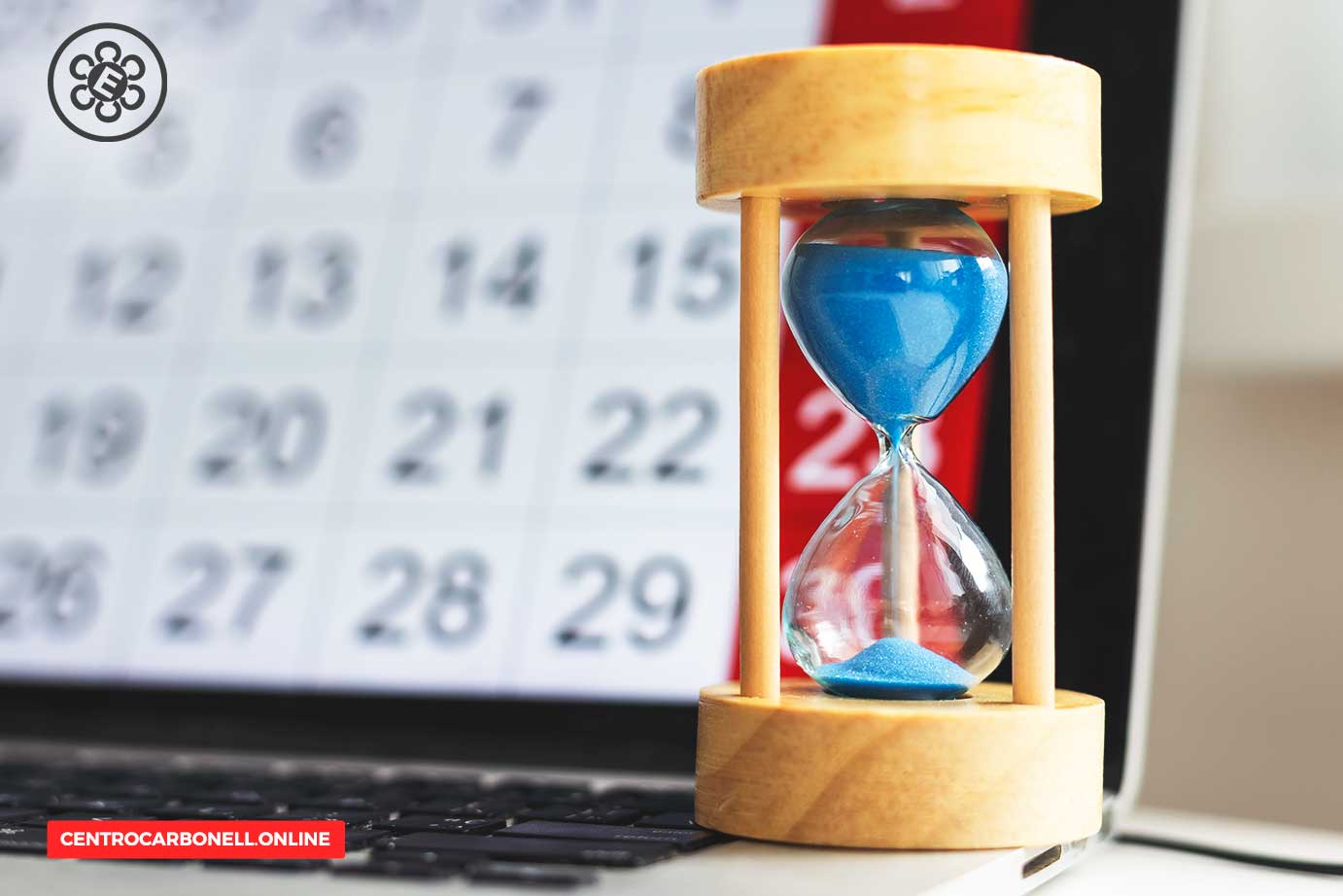 Reloj de arena con arena azul sobre un teclado de computadora, con un calendario desenfocado al fondo.