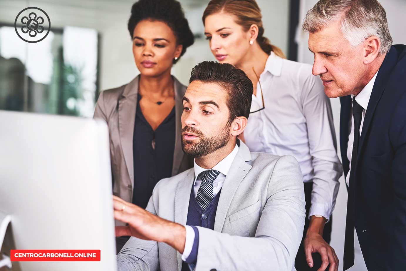Grupo de cuatro personas en un entorno de oficina, observando la pantalla de una computadora. Un hombre vestido de traje gris está concentrado en la pantalla mientras los demás prestan atención.