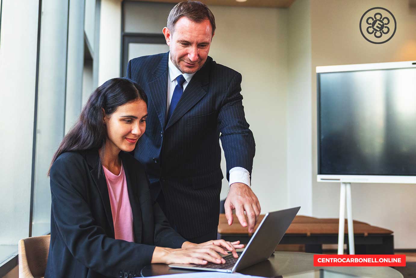 Un hombre de negocios en traje ayuda a una mujer sentada frente a una laptop en una oficina moderna.