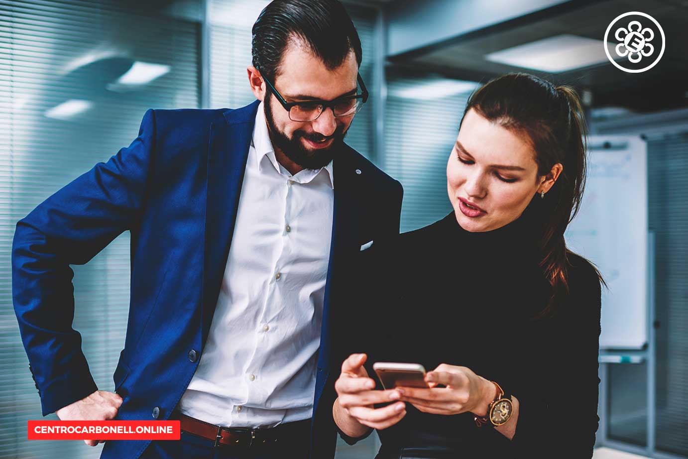Un hombre de negocios con traje azul observa a una mujer que sostiene un teléfono móvil, ambos conversando en un entorno de oficina moderna.