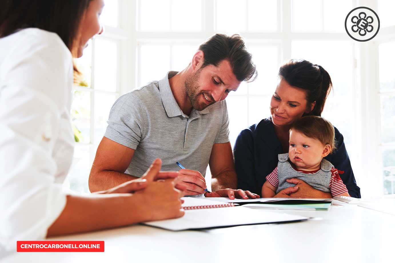 Familia sentada en una mesa, firmando documentos. Un hombre escribe en un cuaderno mientras una mujer lo observa con un bebé en brazos.